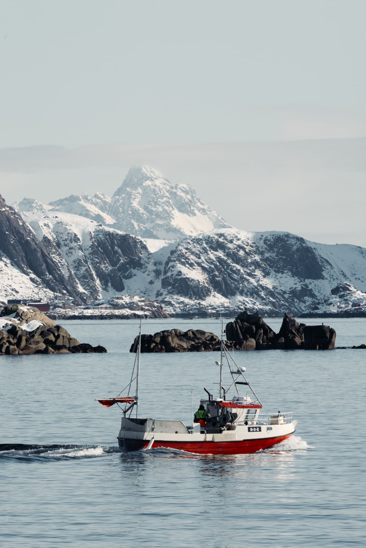A small red coastal fishing boat heading out across a fjord, snow-covered mountains rising behind